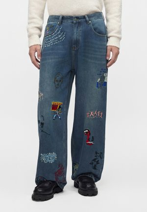 Personne portant un jean bleu ample avec des patchs brodés colorés et des chaussures noires, debout devant un fond blanc uni.