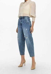 Jasno beżowa bluzka w kropki z bufkami, w zestawie z wysokim stanem, zniszczonymi niebieskimi dżinsami i cielistymi szpilkami.