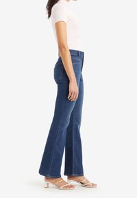 Mujer con jeans acampanados de talle alto azules, top blanco de manga corta y sandalias blancas de tacón, de pie de perfil sobre fondo blanco.