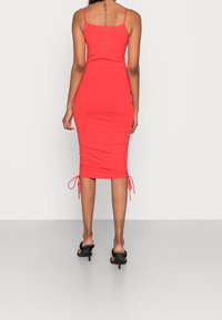 Robe bodycon rouge avec fines bretelles, détails froncés sur les côtés et liens ajustables à l'ourlet. Fabriquée en tissu lisse et élastique.