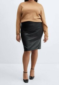 Femme portant un pull à manches longues camel, une jupe crayon en cuir noir et des talons à bride chevillière noirs, debout sur un fond blanc.