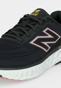 Μαύρο αθλητικό παπούτσι New Balance με ροζ λογότυπο, μαύρα κορδόνια, κίτρινη σήμανση στη γλώσσα και υφασμένο λευκό πέλμα με ροζ λεπτομέρειες.
