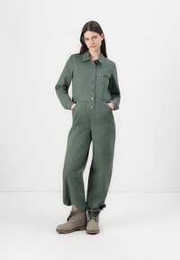 Combinaison vert olive à l'avant avec boutons, poches poitrine et manches longues. Associée à des bottes robustes à lacets brun clair.