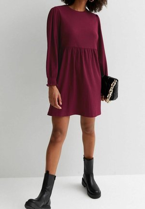 Frau trägt ein bordeauxrotes Mini-Kleid mit langen Ärmeln, schwarze Stiefel bis zur Mitte der Waden und hält eine schwarze gesteppte Clutch mit einer goldenen Kette.