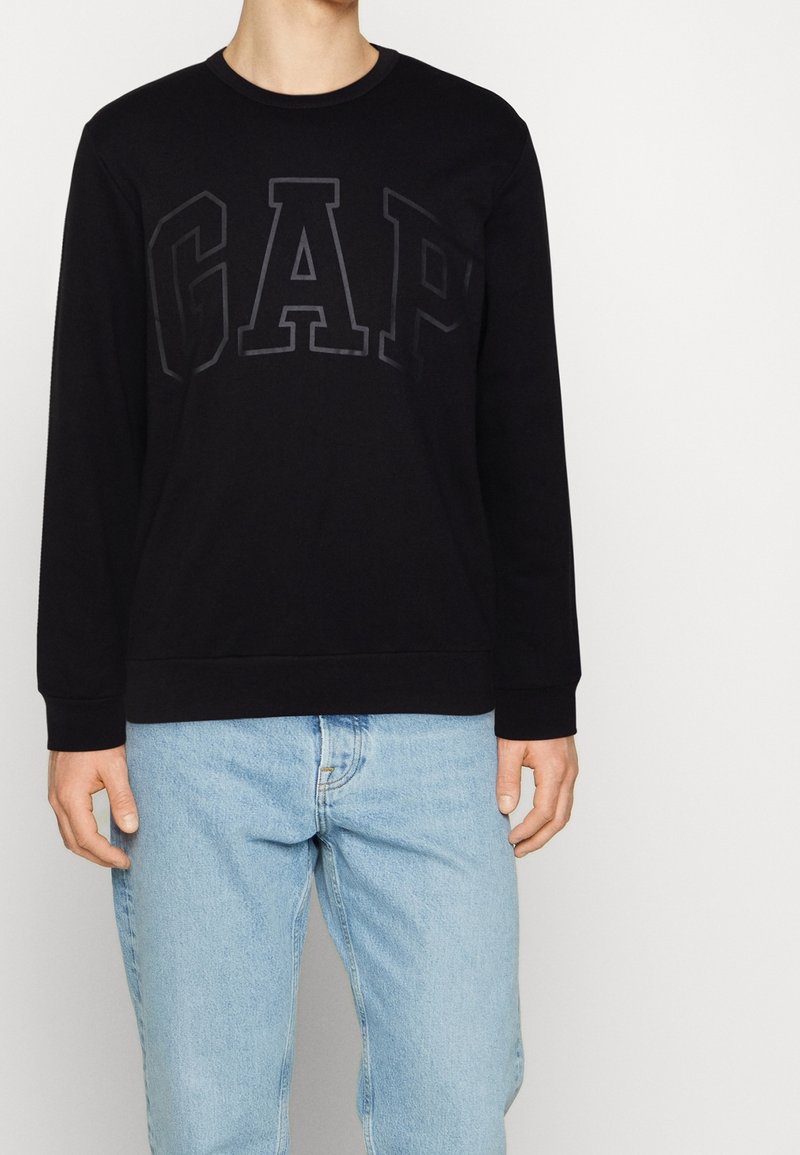 Personne portant un sweat-shirt noir GAP avec un logo en relief foncé et un jean en denim bleu clair, debout devant un fond blanc.