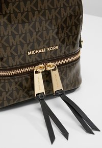 Sac Michael Kors marron avec motif MK répété, tirettes de fermeture éclair dorées gravées du nom de la marque et pampilles en cuir noir sur les fermetures éclair.