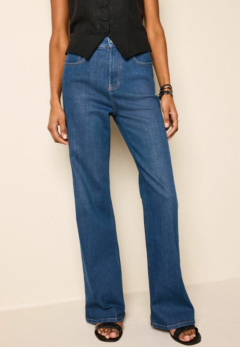Personne portant un jean bleu taille haute à jambes droites, un top noir sans manches, des sandales noires et des bracelets au poignet droit.