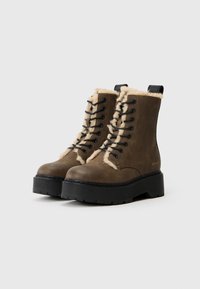 Botas de tobillo de cuero marrón con forro de piel sintética, cierre de cordones negros, suela de plataforma de goma texturizada y tirador en la parte posterior.