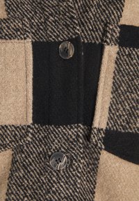 Ylleplädjacka med ett beige och svart rutmönster, med stora knappar och en mjuk textur. Inkluderar svarta detaljer.