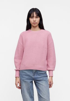 Ung kvinde med langt sort hår iført en lyserød striktrøje og blå jeans, stående mod en ensfarvet hvid baggrund.