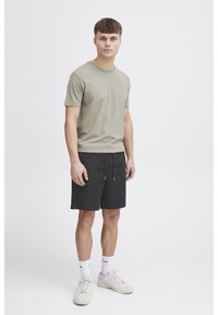 Hele oliiviroheline puuvillane T-särk, mida on kombineeritud mustade päraga pükstega. Modell kannab valgeid sokke ja kreemikaid vabaajajalatseid, seisab neutraalsel taustal.