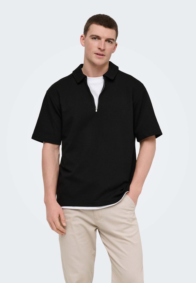 Jauns vīrietis, kurš valkā melnu krekli ar īsām piedurknēm un rāvējslēdzēju pār baltu T-kreklu, kopā ar gaiši bēšām biksēm, stāvot ar vienu roku kabatā.