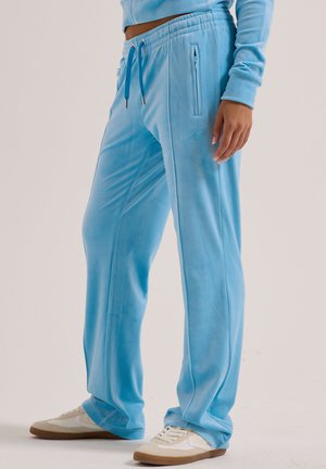 Osoba w jasno niebieskich welurowych spodniach dresowych z zamykanymi na zamek bocznymi kieszeniami i białych trampkach stojąca na jednolitym tle.