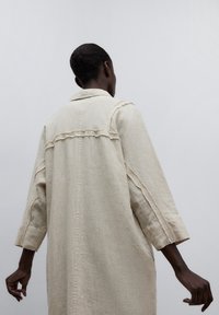 Cappotto in lino beige chiaro con vestibilità ampia, caratterizzato da un orlo grezzo, cuciture a vista e colletto. Maniche lunghe e silhouette dritta.