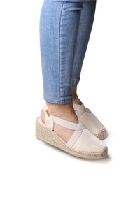 Beige wedge sandals with crisscross elastic straps, jute-wrapped soles, and a smooth canvas upper. Foot partially visible, worn with light blue jeans.