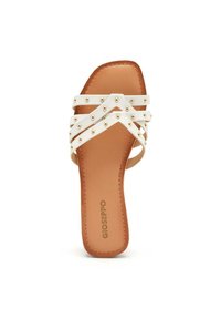 Infradito bianche con cinturini incrociati decorati con borchie dorate, poggiati su una suola beige. Presentano un design piatto e una texture liscia.