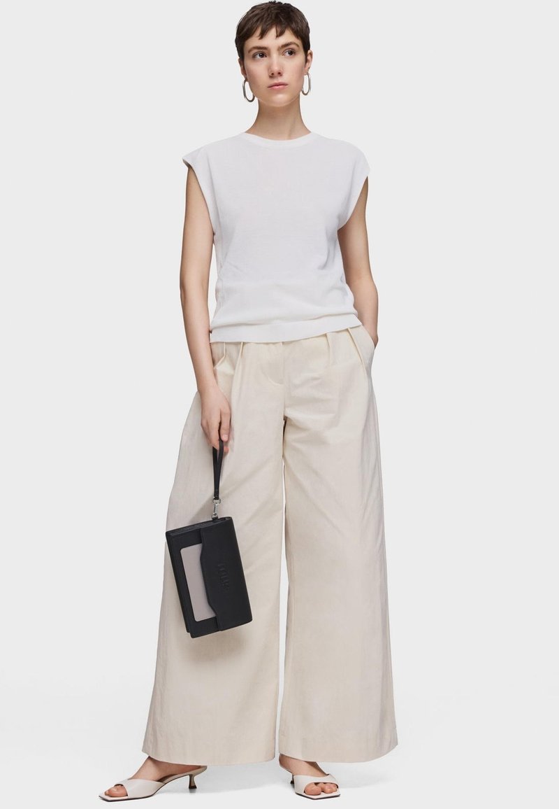 White short-sleeve top paired with cream wide-leg trousers. Black handbag held with a strap. Light-colored sandals. Smooth textures.
