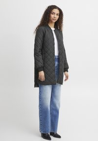 Gepolsterter, dunkelgrüner gesteppter Mantel mit Reißverschluss, gerippten Bündchen und seitlichen Schlitzen, kombiniert mit hellblauen weiten Jeans und schwarzen Stiefeletten.