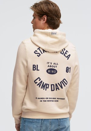Cremefarbener Hoodie aus weichem Stoff, mit großem dunkelblauem Text und Grafiken auf der Rückseite, darunter "CAMP DAVID" und dekorative Elemente.