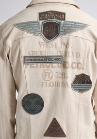 Chaqueta beige con varios parches bordados. Los detalles notables incluyen diseños de alas, logotipos y texto en tonos azul oscuro y marrón.