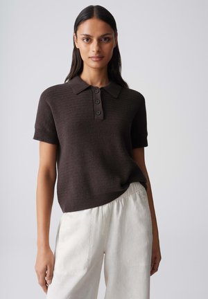 Woman with long dark hair wearing a dark brown short-sleeve knit polo and loose white pants against a plain light background.