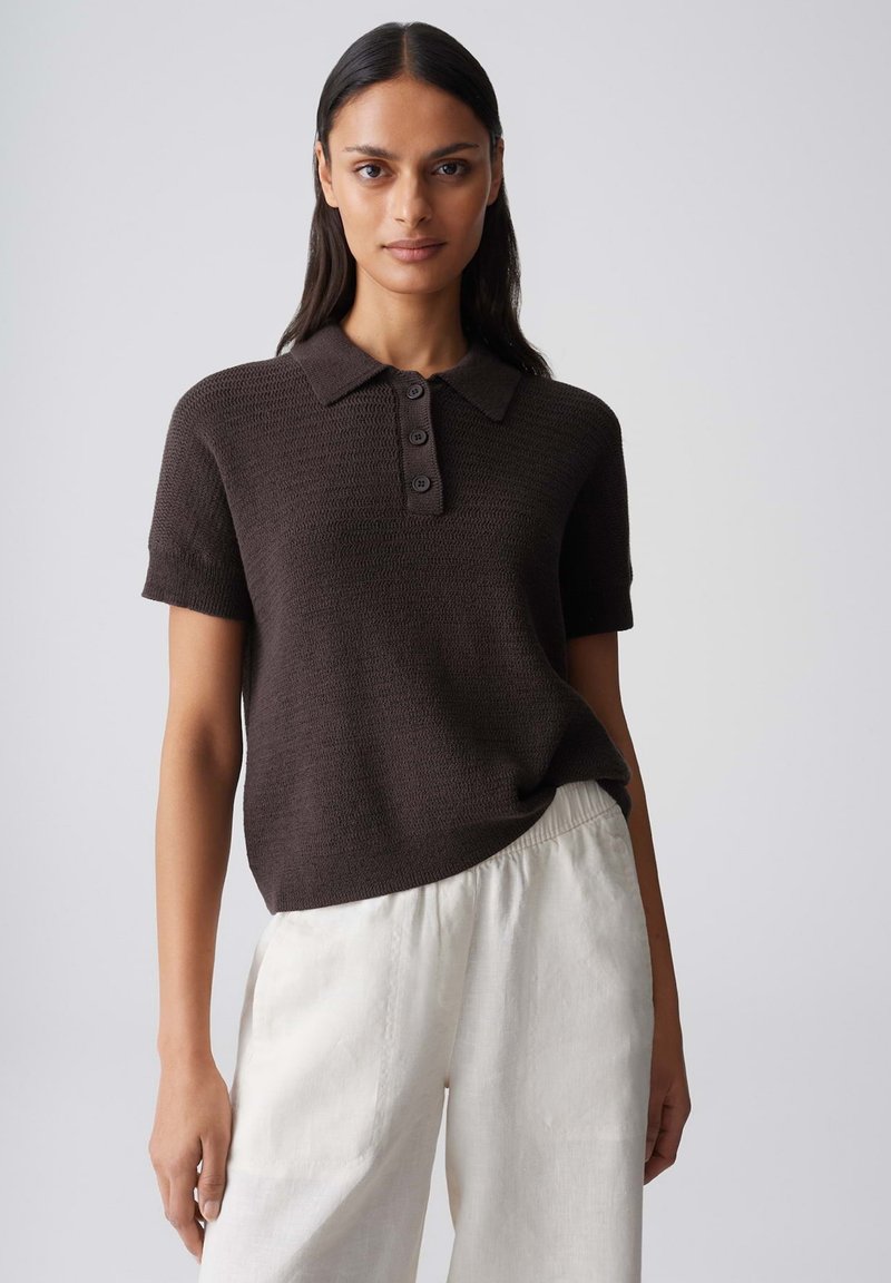 Woman with long dark hair wearing a dark brown short-sleeve knit polo and loose white pants against a plain light background.