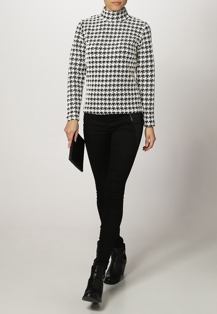 Femme portant un col roulé pied-de-poule noir et blanc, pantalon noir, bottines noires, tenant une pochette noire, marchant vers l'avant sur un fond uni.