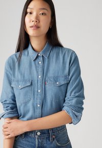 Lyseblå denimskjorte med knapp-oppsett, to brystlommer og oppbrettede ermer som har metallknapper og en subtil tekstur.