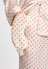 Máscara de sueño de seda con forma redondeada, fondo crema y estampado de corazones rojos dispersos. La máscara cuenta con un lado fruncido para un ajuste ajustable.
