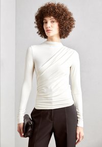 Langærmet off-white top med rynket detaljering og rund halsudskæring, parret med mørke bukser og en sort quiltet clutch.