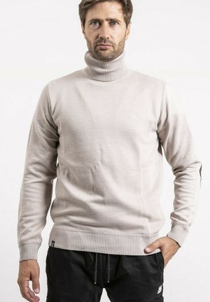 Homme portant un pull à col roulé beige et un pantalon noir, debout devant un fond blanc uni, main dans la poche.