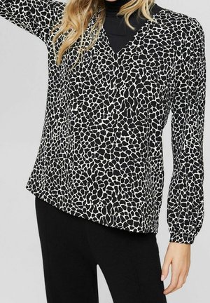 Mujer vestida con una blusa de manga larga con un patrón abstracto en blanco y negro sobre un cuello alto negro y pantalones negros.