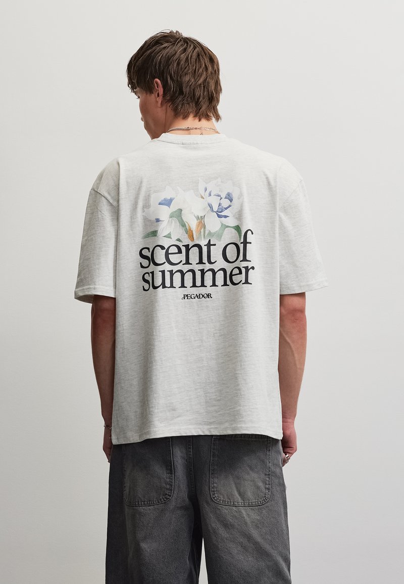 Νεαρός άντρας που φοράει ανοιχτογόρ T-shirt με floral σχέδιο και το κείμενο «scent of summer» στην πλάτη, σε συνδυασμό με σκούρο γκρι παντελόνια.