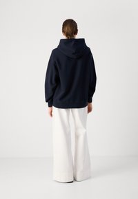 Hoodie oversized azul-marinho com bolso canguru e capuz ajustável, combinado com calças brancas de pernas largas, exibido pela parte de trás.