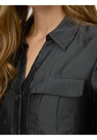 Schwarzes, geripptes Hemd mit Kragen, vorderer Knopfleiste und zwei aufgesetzten Brusttaschen mit Klappen. Glatte Textur.