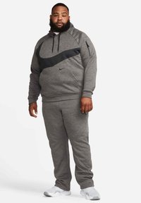 Homme portant un sweat à capuche Nike gris avec un swoosh noir, un pantalon de jogging gris assorti et des baskets blanches, debout devant un fond blanc.