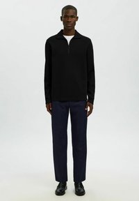 Zwarte zip-hals pullover, lange mouwen, gestructureerde stof; gecombineerd met marineblauwe broek en zwarte leren schoenen, staand voor een lichte achtergrond.
