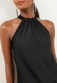Čierny top na zaväzovanie za krk s nazbieraným výstrihom, jemnou látkou a hladkou textúrou. Dopĺňajú ho veľké zlaté a čierne náušnice.
