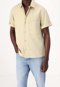 Lichtbeige kortemouwenshirt met een gestructureerde afwerking, een borstzak en knoopsluiting, gecombineerd met lichtblauwe spijkerbroek.