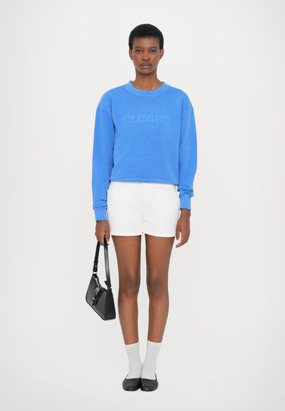 Μπλε φούτερ με το κείμενο "CLOSED", λευκά σορτς, γκρι κάλτσες και μαύρες μπαλαρίνες. Το μοντέλο κρατά μια μαύρη τσάντα, στέκεται σε ένα ο�υδέτερο φόντο.