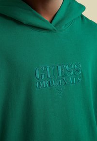 Groene katoenen hoodie met een geborduurd "GUESS ORIGINALS" logo in een bijpassende kleur. De stof heeft een zachte textuur en een ontspannen ontwerp.