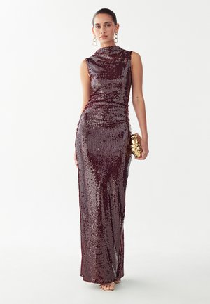 Mujer con un vestido sin mangas con lentejuelas burdeos hasta el suelo, sosteniendo un bolso de mano dorado con textura y llevando grandes pendientes de aro.