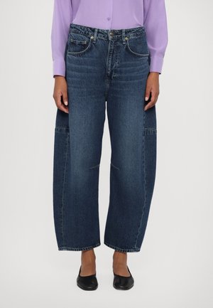 Person iført brede mørkeblå jeans, sorte flade sko og en langærmet lysegrå/lilla knap-på-skjorte, stående foran en ensfarvet baggrund.