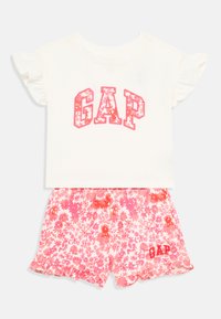 Dvoudílný outfit skládající se z bílého topu s krátkým rukávem, na kterém je růžový květinový potisk "GAP", a růžových květinových šortek s volánkovým lemem a logem.