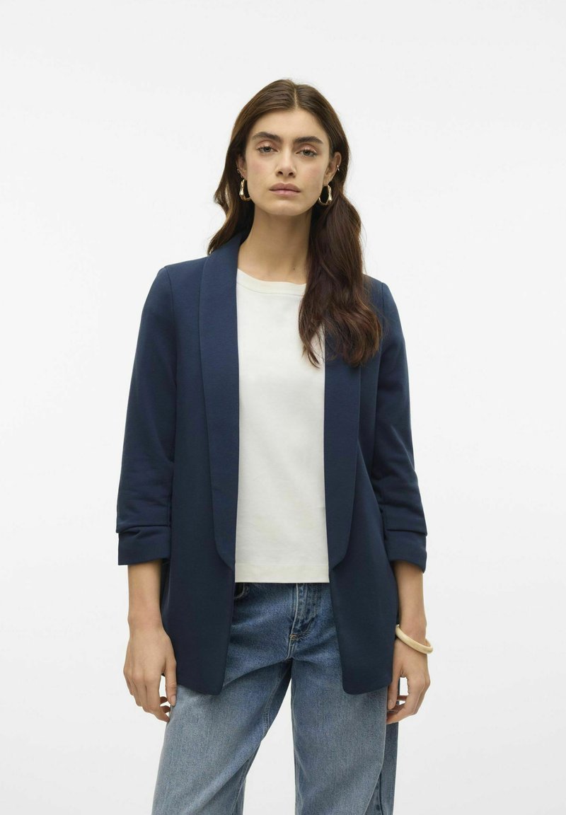 Blazer marine à la texture lisse, à l'avant ouvert et aux manches retroussées, associé à un t-shirt blanc et à un jean bleu clair. Design simple.