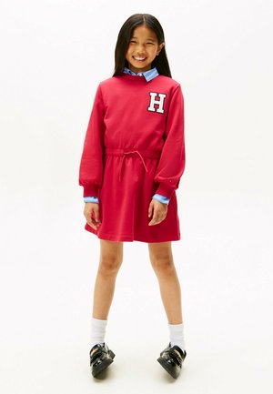 Rode sweatshirtjurk met een trekkoord in de taille, met een witte "H" logo. Lange mouwen, gelaagd over een blauwe overhemd met kraag, gecombineerd met zwarte schoenen.
