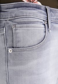 Jeans de mezclilla gris claro que presentan un bolsillo trasero con herrajes plateados. La tela tiene una textura suave con un ligero desvanecimiento.