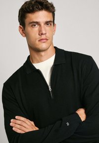 Schwarzer Zip-Pullover mit Kragen, gerippten Bündchen und einem kleinen Logodetail am Ärmel. Weicher Strickstoff, minimales Design.