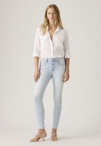 Γυναίκα με μακριά ξανθά μαλλιά που φοράει λευκό πουκάμισο με κουμπιά, ανοιχτόχρωμα στενά τζιν και μπεζ σανδάλια με λουράκια και χαμηλό τακούνι, όρθια.