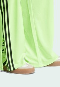 Limoengroene sportbroek met zwarte strepen aan de zijkanten, gecombineerd met grijze en witte schoenen en patronen in de sokken die zichtbaar zijn bij de ton.